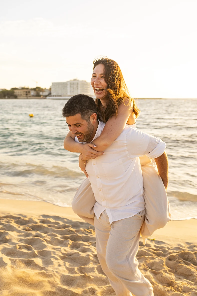 Couple laughing as he carries her on his back by the sea during a family photography session in Mallorca