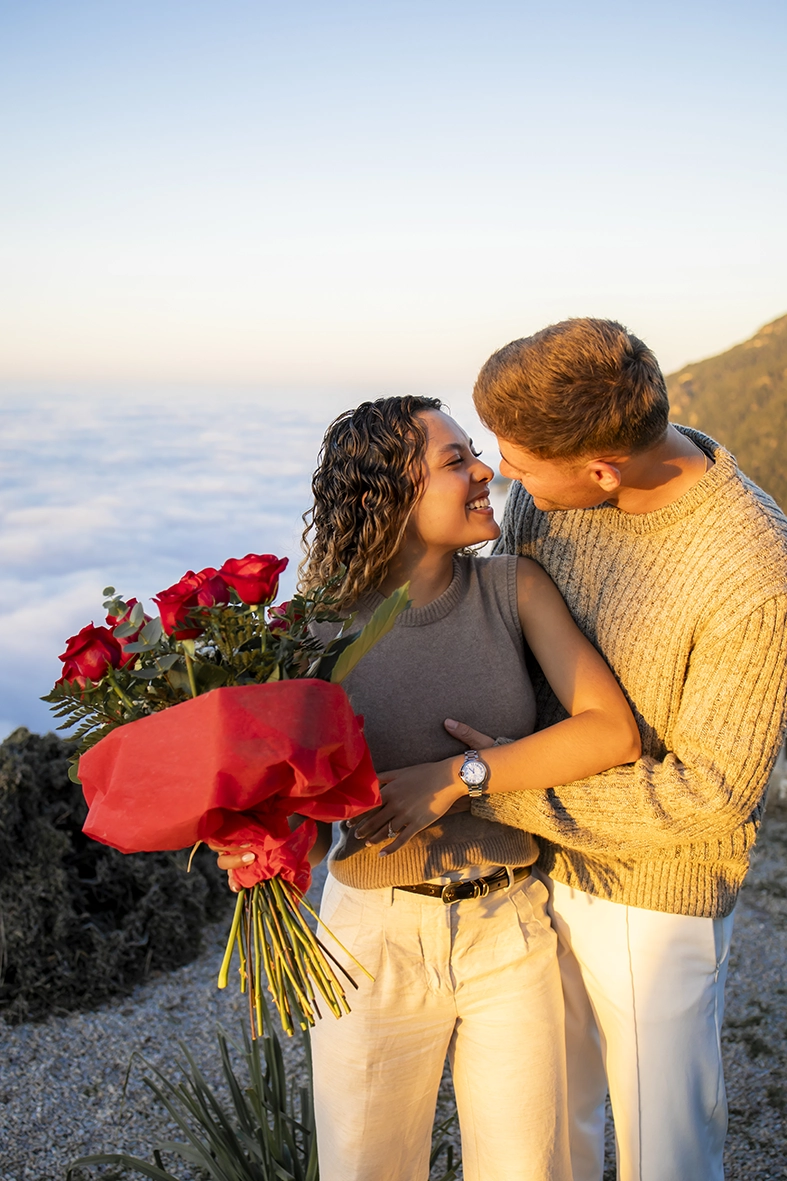 Couple embracing with red rose bouquet after exclusive marriage proposal in Mallorca on cliffs above sea of clouds.
