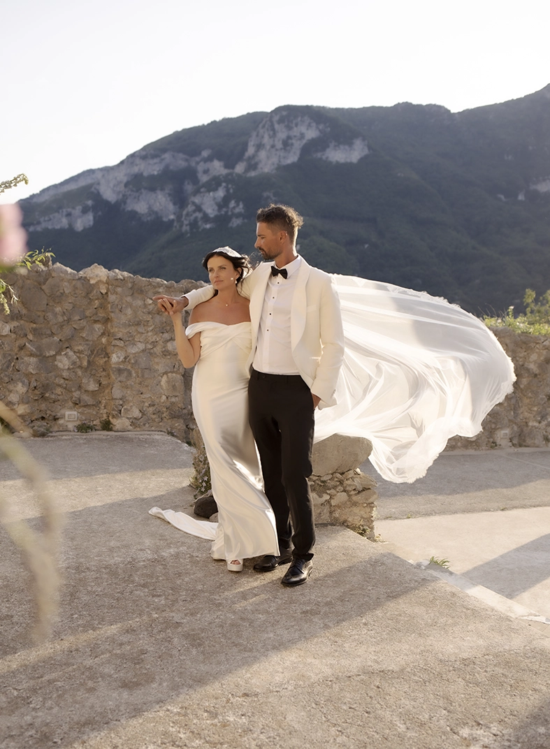 Novios posando con el velo al viento y las montañas de Ravello al fondo durante su boda en la Costa Amalfitana.