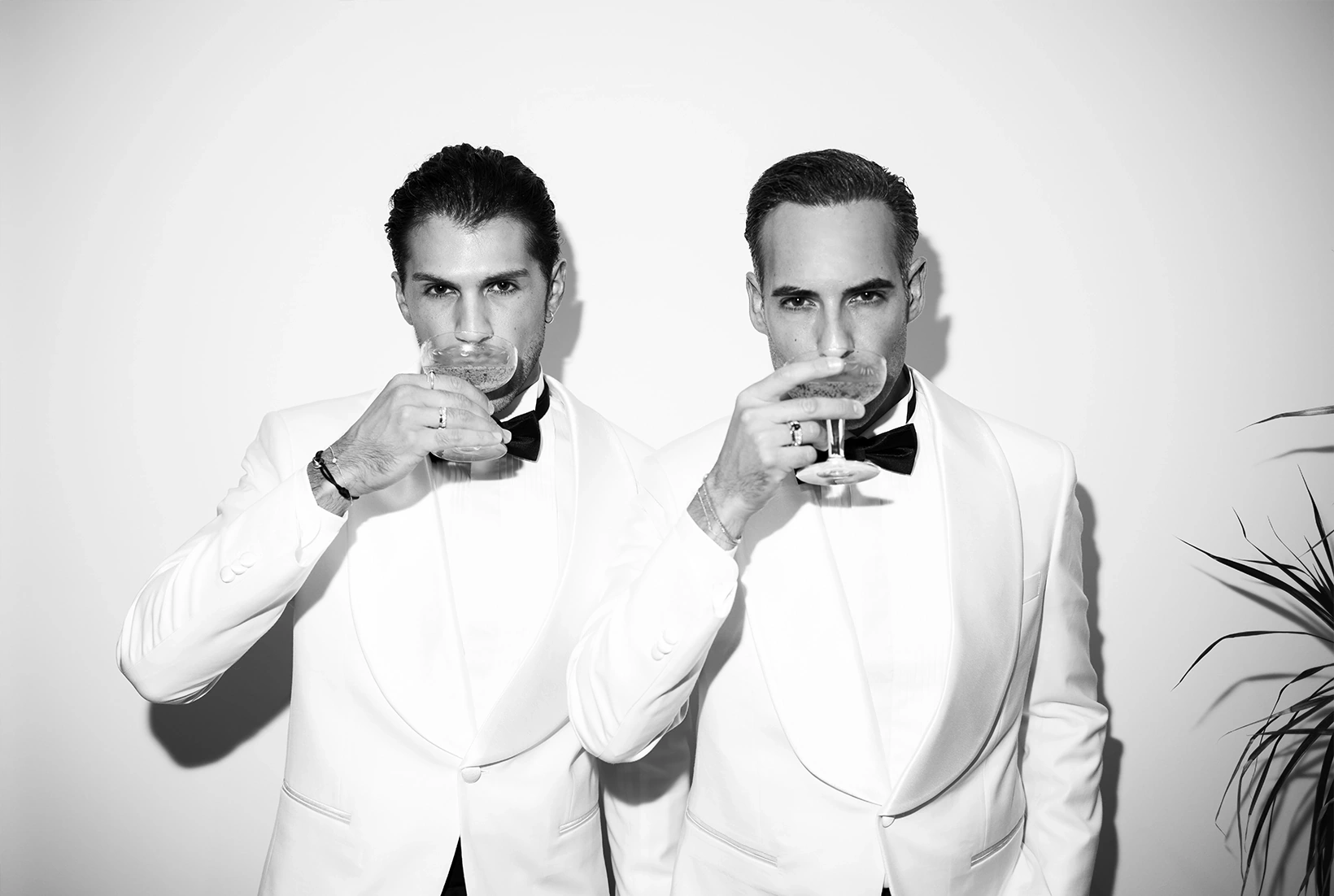 Two grooms in white tuxedos sharing a drink during an elegant wedding in Mallorca.