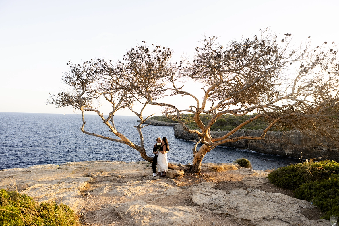 Pareja besándose junto a un árbol frente al mar en uno de los mejores lugares para pedida en Mallorca. Fotografía de Inma del Valle.