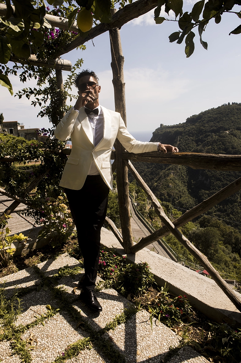 Groom in a white tuxedo enjoying a cigar beside a terrace overlooking the Amalfi Coast.