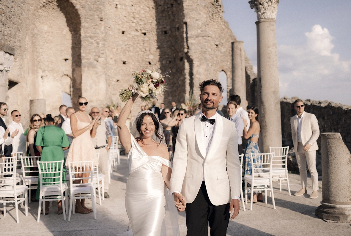 Newlyweds walking hand in hand as the bride raises her bouquet after their wedding ceremony in Scala.